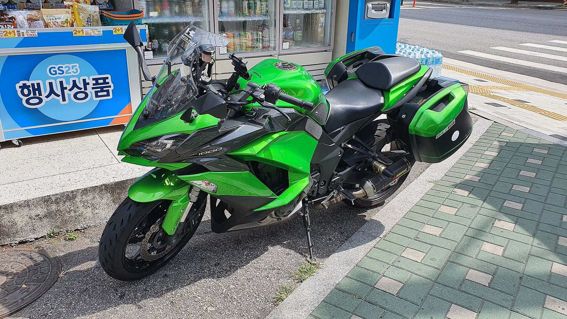 A green Kawasaki road motorcycle fitted with hard panniers, parked at a kerb