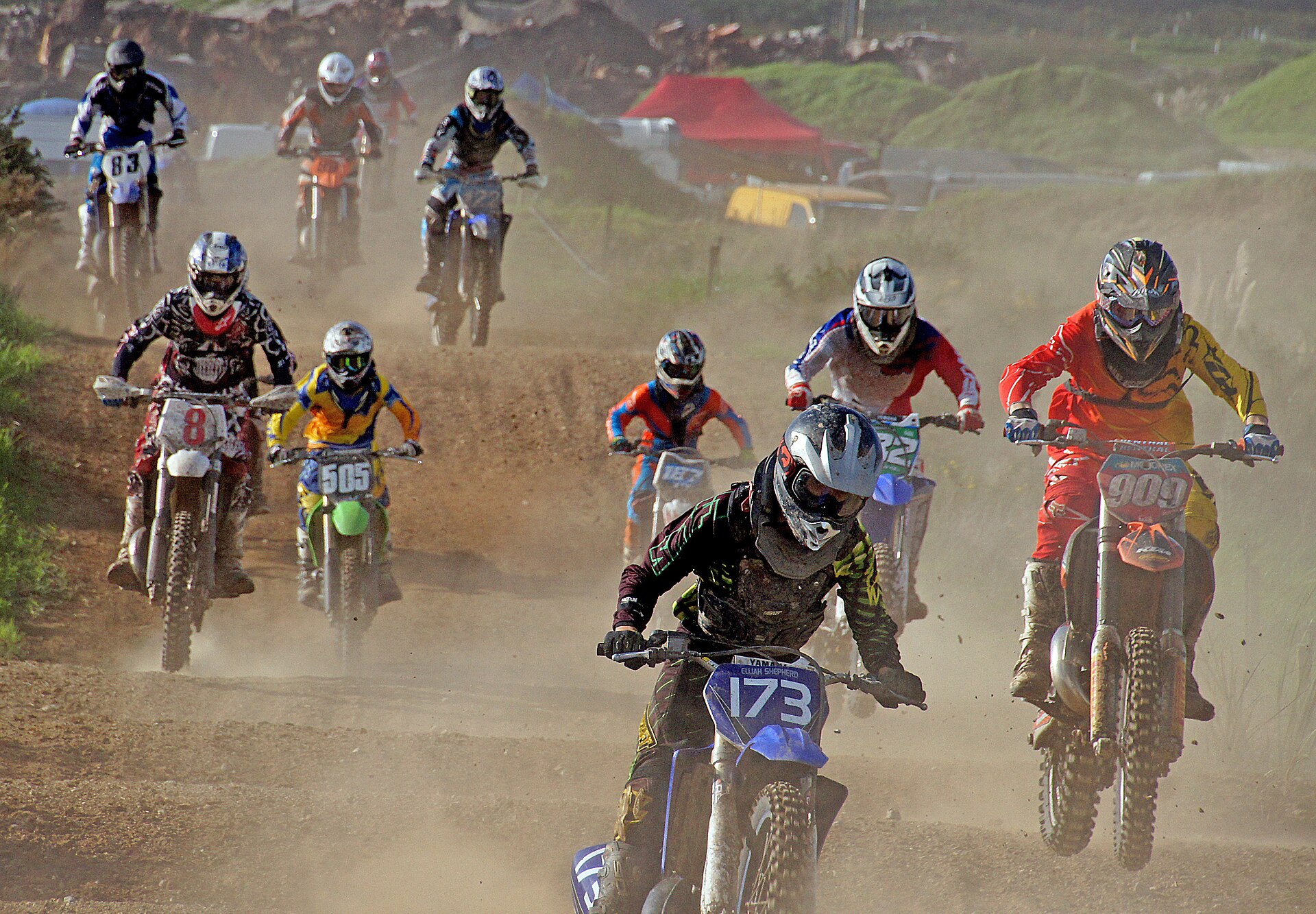 A pack of motocross riders in full helmets, boots and body armour accelerating through dust